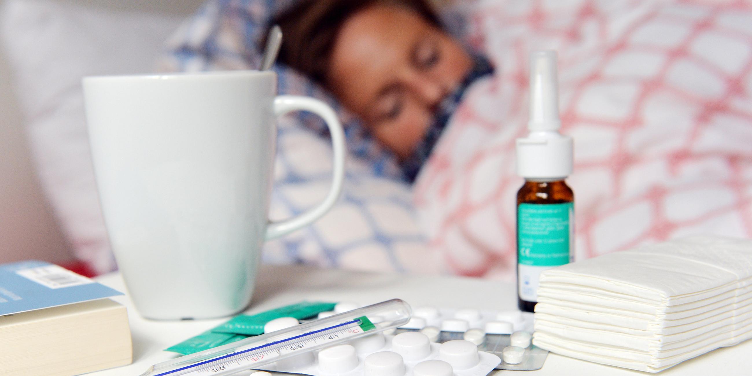 Eine erkältete Person liegt im Bett, auf einem Nachttisch liegen Medikamente und ein Fieberthermometer.