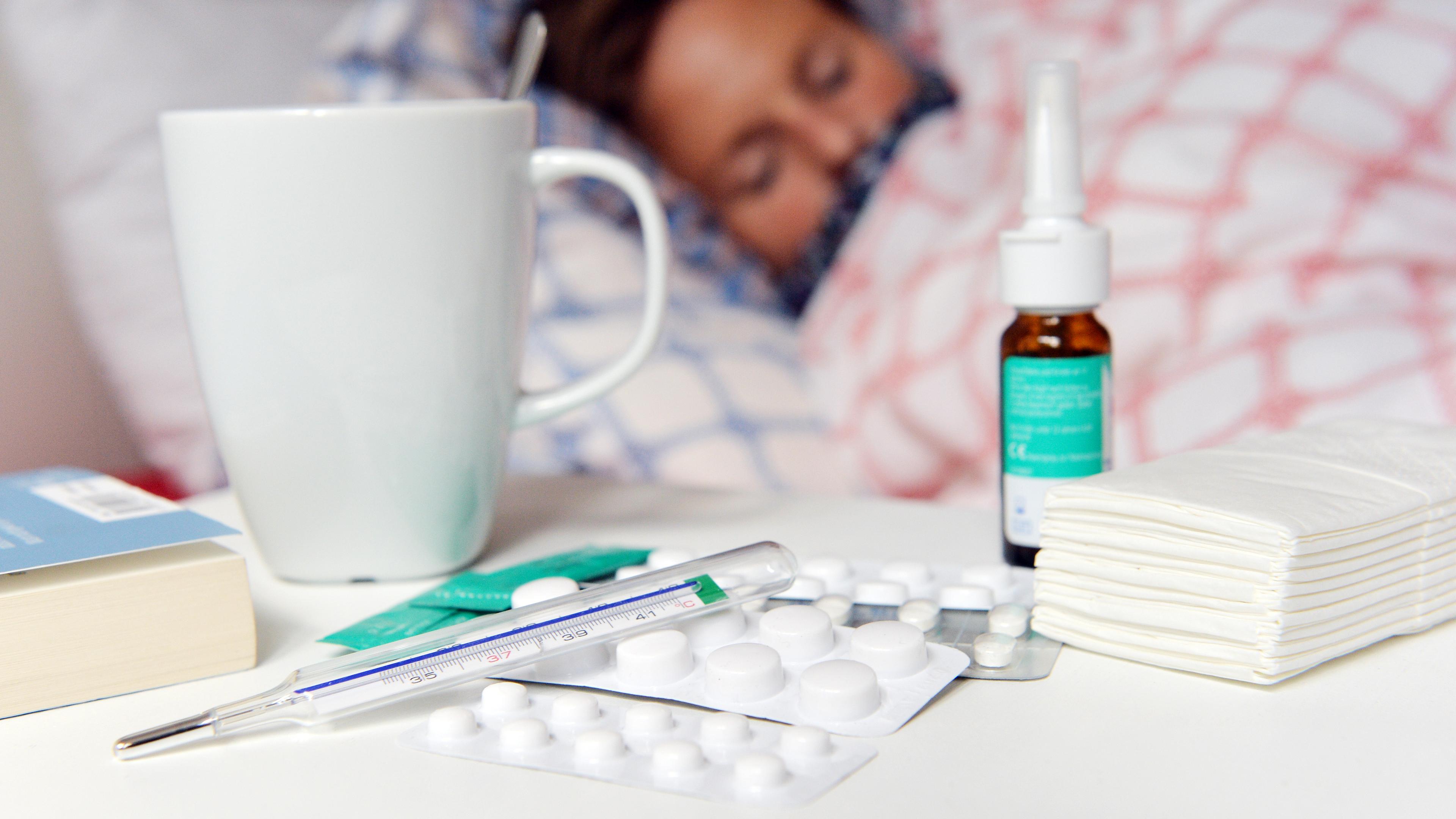 Eine kranke Person liegt im Bett, vor ihr eine Reihe Medikamente und ein Fieberthermometer. (Symbolbild)