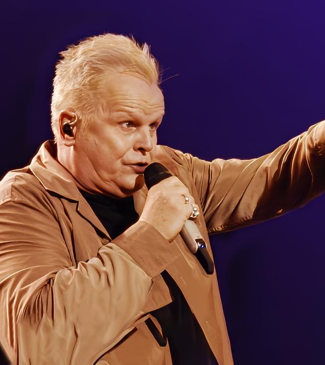 Herbert Grönemeyer singend mit Mikrofon vor dem Mund, den linken Arm mit Zeigefinger deutend zum Publikum. Er trägt einen Mantel in beige.