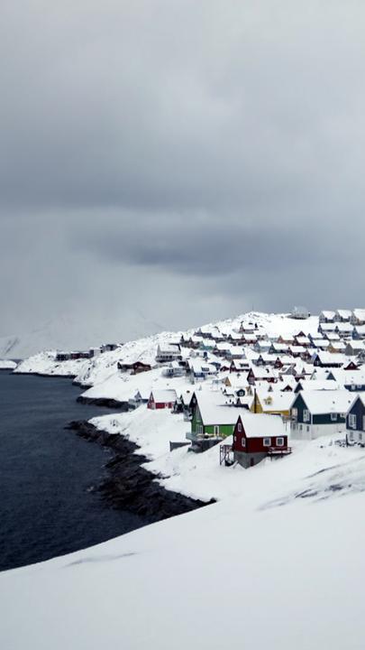 Zu sehen ist eine verschneite Küstenregion in Grönland: links das Meer, rechts Häuser.