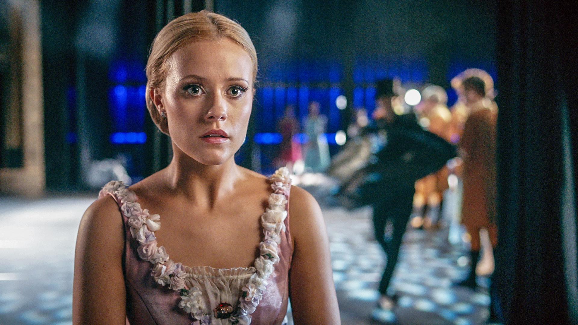 Lena trägt das Kostüm der Libertalia. Sie steht auf der Bühne des Grand Prix, im Hintergrund rechts sind unscharf ihre Mitschüler zu erkennen. Lena sieht geschockt aus.