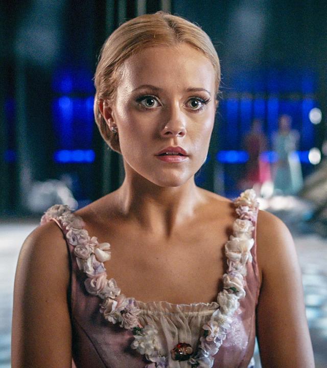 Lena trägt das Kostüm der Libertalia. Sie steht auf der Bühne des Grand Prix, im Hintergrund rechts sind unscharf ihre Mitschüler zu erkennen. Lena sieht geschockt aus.