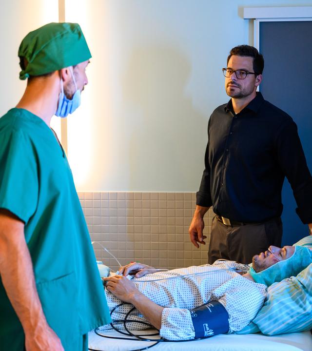 "Bettys Diagnose - Großer Bruder": Dr. Koopmann (Tobias Licht) steht neben dem Krankenhausbett seines Bruder (Tobias van Dieken). Dieser liegt vorbereitet für die anstehende OP bereits in Narkose. Dr. Frank Stern, steht in OP Kleidung auf der anderen Seite des Bettes und schaut Dr. Koopmann an.