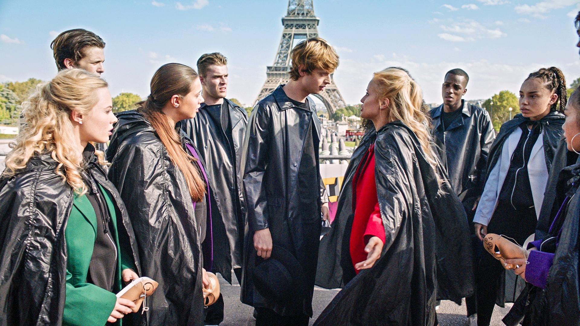 Lena (Mitte rechts) und Jeff (Mitte links) diskutieren miteinander. Um sie herum stehen andere Tänzer und Tänzerinnen, alle tragen lange, schwarze Mäntel. Im Hintergrund steht der Eiffelturm.