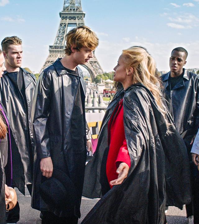 Lena (Mitte rechts) und Jeff (Mitte links) diskutieren miteinander. Um sie herum stehen andere Tänzer und Tänzerinnen, alle tragen lange, schwarze Mäntel. Im Hintergrund steht der Eiffelturm.