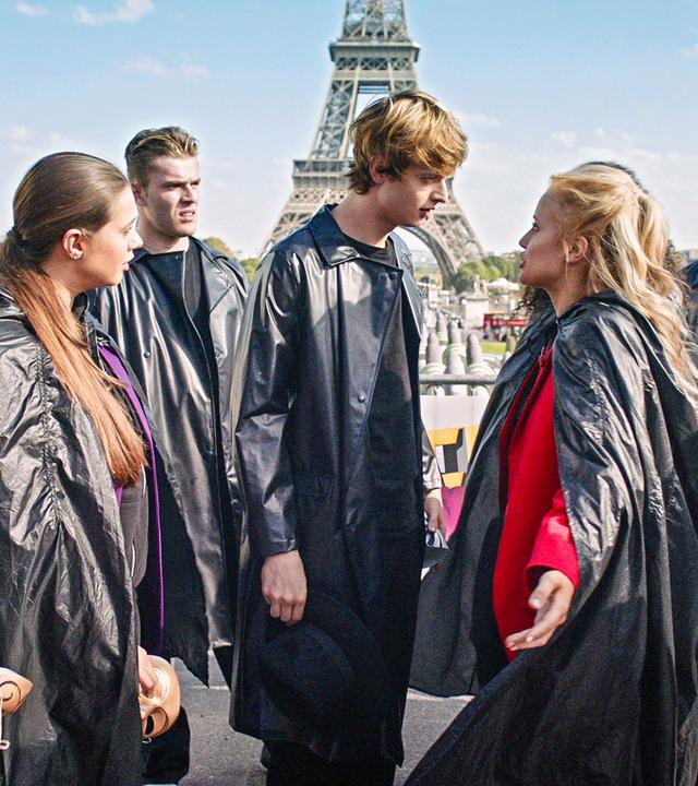 Lena und Jeff diskutieren miteinander. Um sie herum stehen andere Tänzer und Tänzerinnen, alle tragen lange, schwarze Mäntel. Im Hintergrund steht der Eiffelturm.