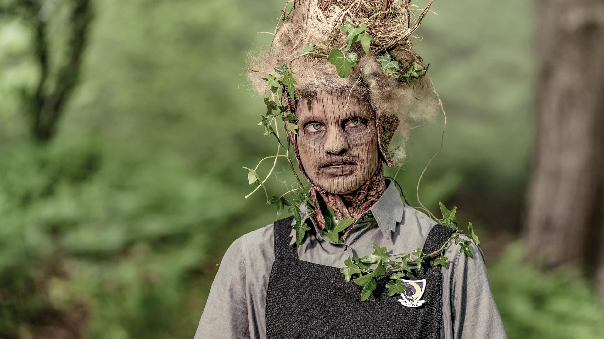 Tagsüber in einem Waldstück: Esther verwandelt sich mehr und mehr in einen Baum. Ihr Gesicht hat Ähnlichkeit mit einem Baumstamm, auf ihrem Kopf und an den Armen wachsen Blätter.