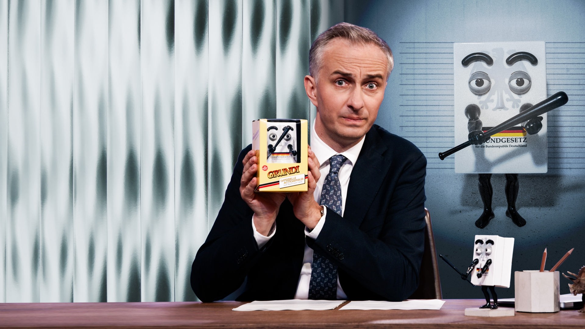 Jan Böhmermann sitzt am Schreibtisch und hält eine "ZDF Magazin Royale"-Actionfigur "Grundi" in den Händen. Rechts im Hintergrund ist ein Bild von "Grundi" zu sehen.