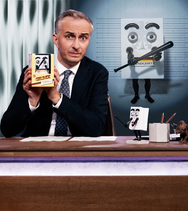 Jan Böhmermann sitzt am Schreibtisch und hält eine "ZDF Magazin Royale"-Actionfigur "Grundi" in den Händen. Rechts im Hintergrund ist ein Bild von "Grundi" zu sehen.