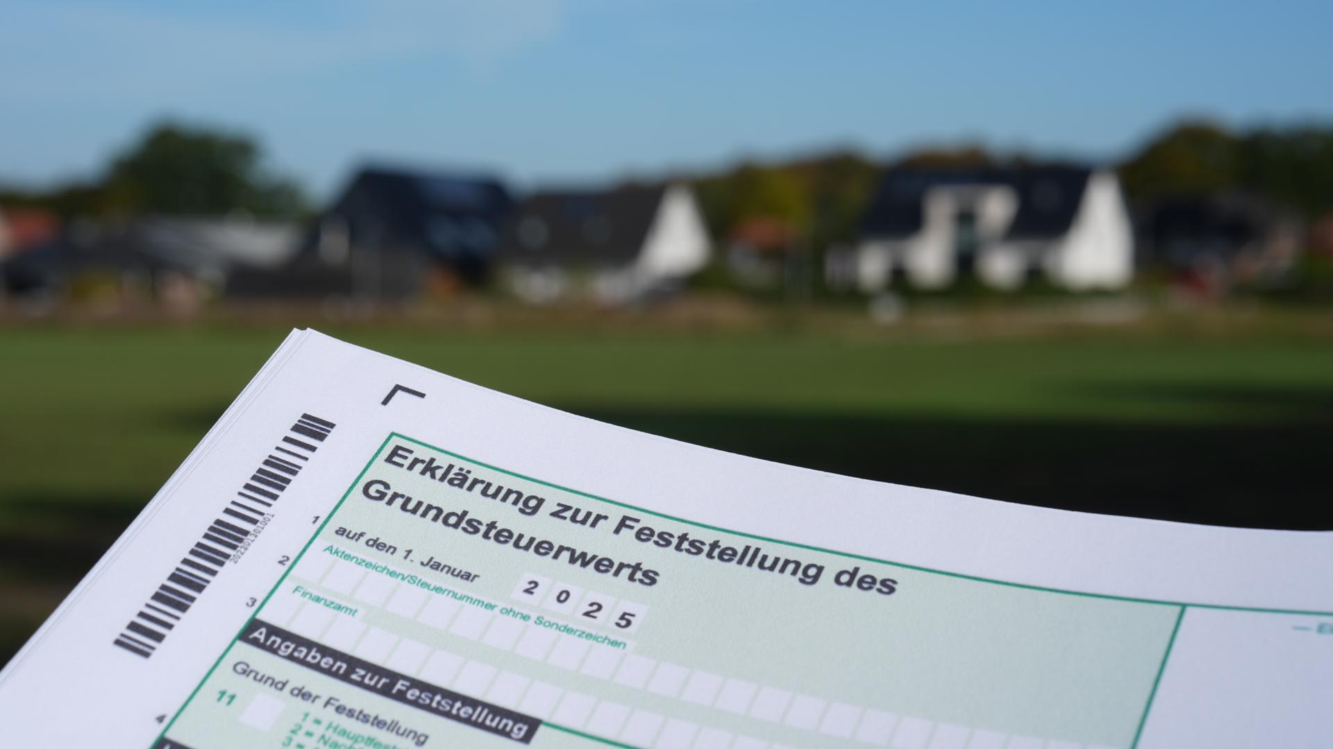 17.10.2024, Schleswig-Holstein, Jersbek: Ein Erklärung zur Feststellung des Grundsteuerwerts für das Jahr 2025 wird vor eine Wohnsiedlung hochgehalten.