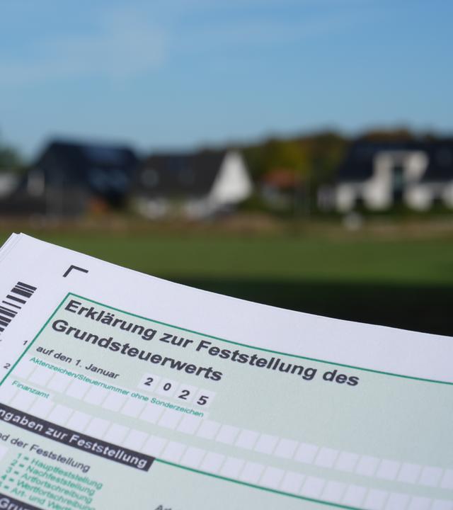 17.10.2024, Schleswig-Holstein, Jersbek: Ein Erklärung zur Feststellung des Grundsteuerwerts für das Jahr 2025 wird vor eine Wohnsiedlung hochgehalten.