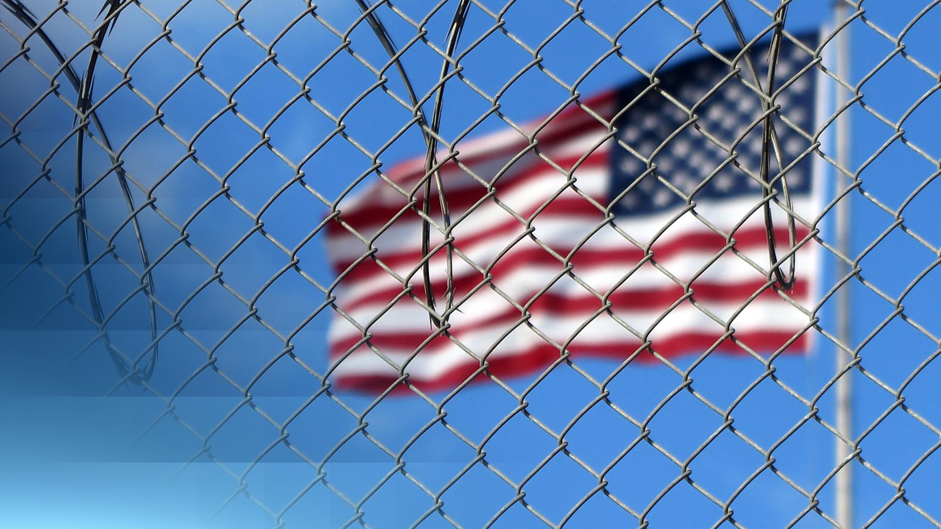 Eine USA-Flagge an einem Mast im Hintergrund leicht unscharf. Davor ein Metallgitterzaun mit sichtbarem Stacheldraht.