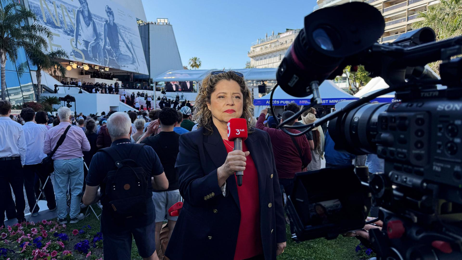 Eine Frau mit einem 3sat-Mikrofon wird von einer Kamera gefilmt, während sie vor dem Kino in Cannes steht und einen Aufsager macht.