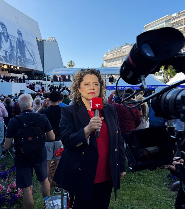 Eine Frau mit einem 3sat-Mikrofon wird von einer Kamera gefilmt, während sie vor dem Kino in Cannes steht und einen Aufsager macht.