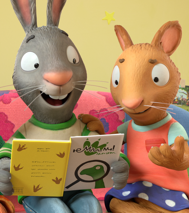 Jamila, das Eichhörnchen, Pip, das Kaninchen und Posy, die Maus, sitzen nebeneinander auf einer pinkfarbenen Couch mit rot-blauen Blumen. Pip hält ein geöffnetes Dino-Buch in beiden Händen.  Alle drei schauen lächelnd in das Buch. Posy hat ihre rechte Hand auf Pips linker Schulter liegen. Ihre linke Hand zeigt geöffnet nach vorne. Neben Jamila liegt ein gelbes Kissen mit grünen Punkten und neben Posy ein grünes Kissen mit Karomuster. An der gelben Wand befinden sich verschiedene Bilder.