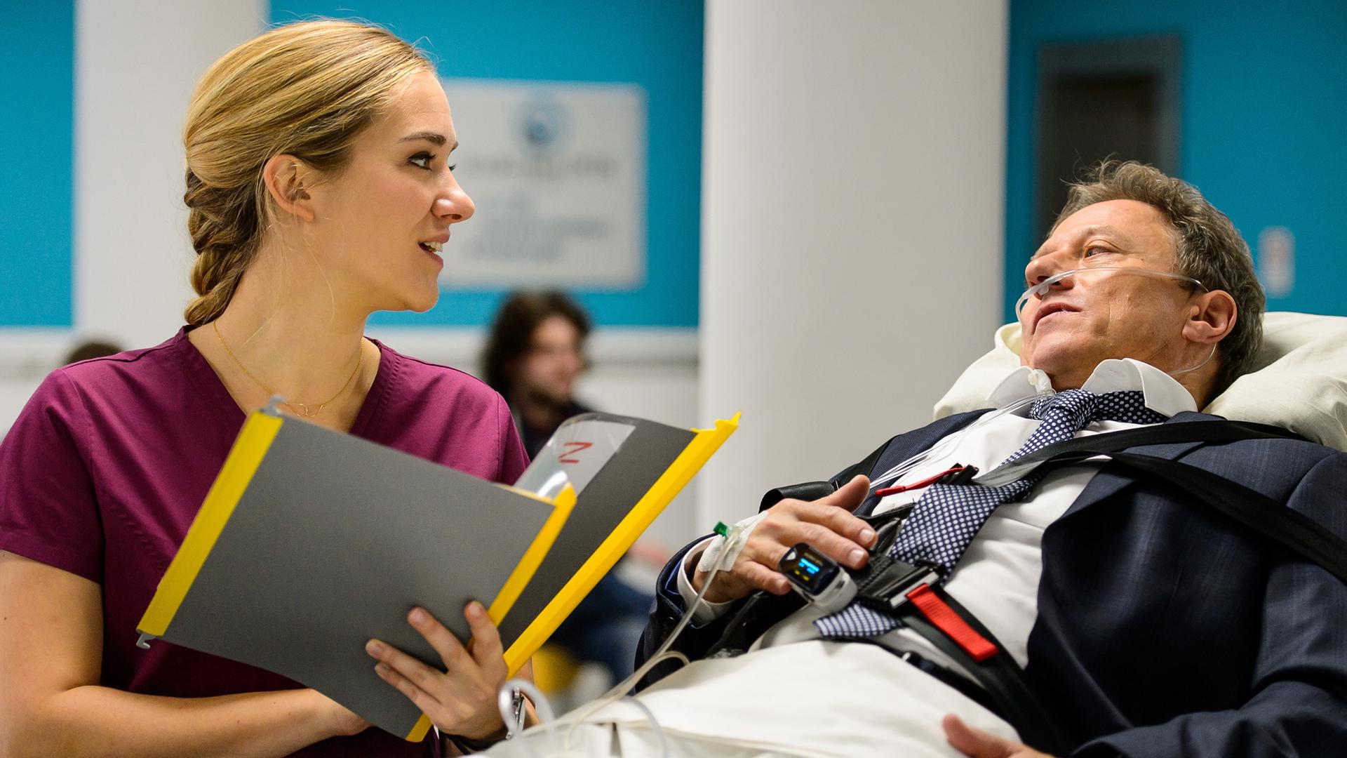 Prof. Friedhelm Lewandowski (Axel Pape) wird auf der Liege in die Karlsklinik eingeliefert. Betty (Annina Hellenthal) steht mit den Papieren in Arm neben ihm
