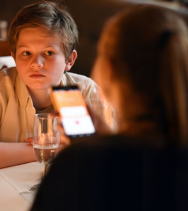 "Frühling - Gute Väter, schlechte Väter": Nicky Söringer (Valentin Wessely) sitzt in einem Restaurant an einem Tisch und blickt erwartungsvoll auf seine Schwester Paula (Lotte Hanné), die auf ihr Handy schaut.