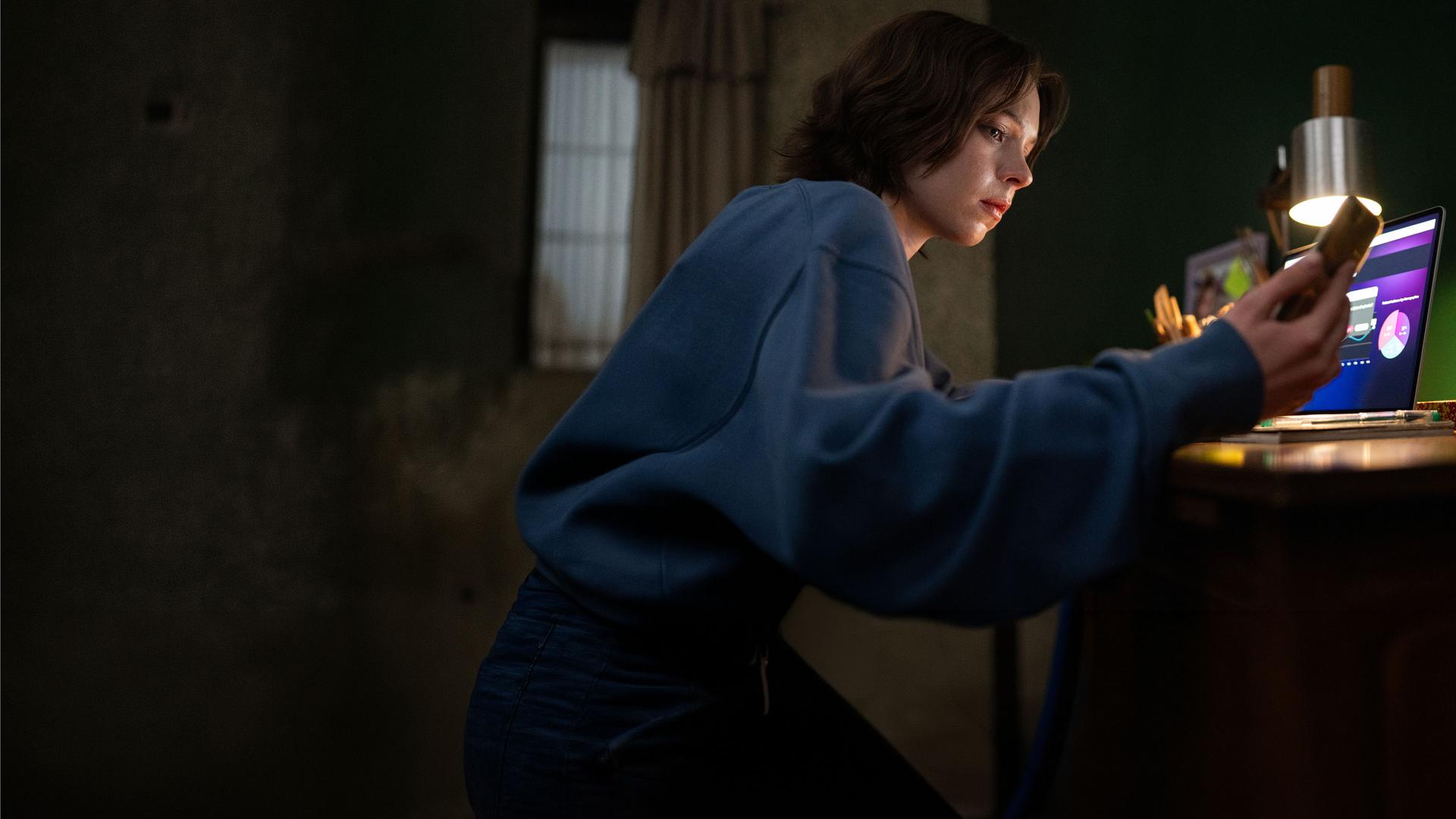 "A Good Girl's Guide to Murder - Gutes Mädchen, böses Blut": Pip Fitz-Amobi (Emma Myers) sitzt im Licht einer Lampe an ihrem Schreibtisch und blickt auf ihr Handy, das sie in der rechten Hand hält. Auf dem Laptop vor ihr sind Graphen zu erkennen.