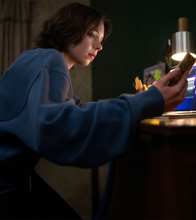 "A Good Girl's Guide to Murder - Gutes Mädchen, böses Blut": Pip Fitz-Amobi (Emma Myers) sitzt im Licht einer Lampe an ihrem Schreibtisch und blickt auf ihr Handy, das sie in der rechten Hand hält. Auf dem Laptop vor ihr sind Graphen zu erkennen.