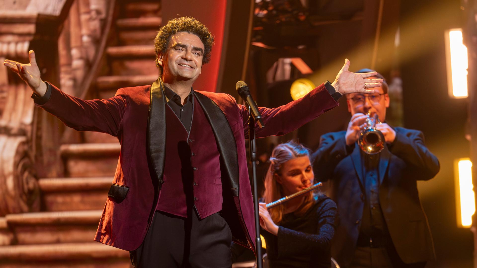"Die Giovanni Zarrella Show – Kleine Stars ganz groß": Rolando Villazón steht mit weit ausgebreiteten Armen vor einem Mikrofon auf der Bühne. Im Hintergrund befinden sich zwei Orchestermusiker mit Trompete und Querflöte.