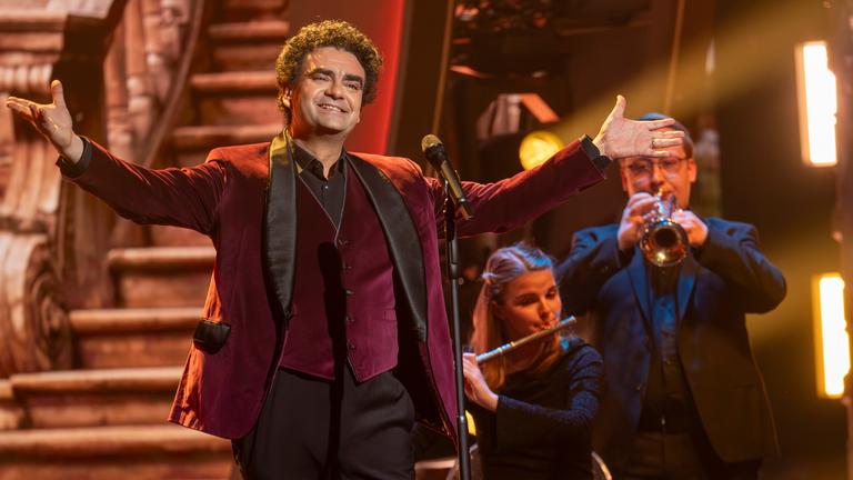"Die Giovanni Zarrella Show – Kleine Stars ganz groß": Rolando Villazón steht mit weit ausgebreiteten Armen vor einem Mikrofon auf der Bühne. Im Hintergrund befinden sich zwei Orchestermusiker mit Trompete und Querflöte.