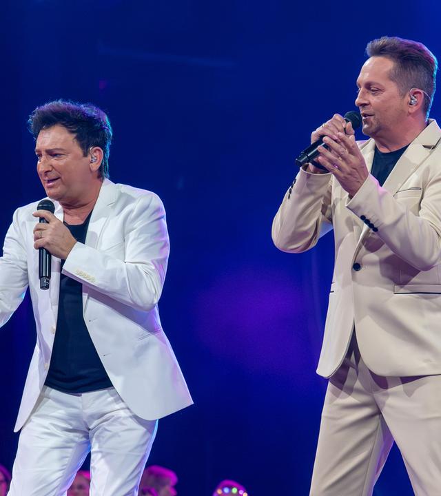 "Die Giovanni Zarrella Show – Kleine Stars ganz groß": Das Schlagerduo Fantasy steht auf der Bühne. Beide tragen helle Anzüge über dunklen T-Shirts, singen in ein Mikrofon und blicken dabei ins Publikum.