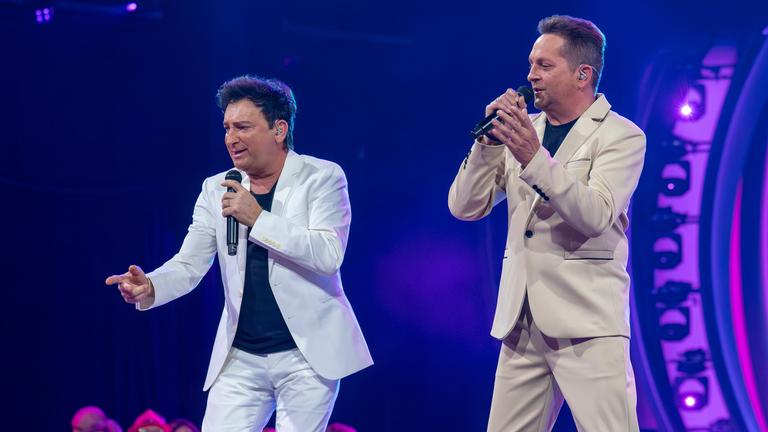 "Die Giovanni Zarrella Show – Kleine Stars ganz groß": Das Schlagerduo Fantasy steht auf der Bühne. Beide tragen helle Anzüge über dunklen T-Shirts, singen in ein Mikrofon und blicken dabei ins Publikum.