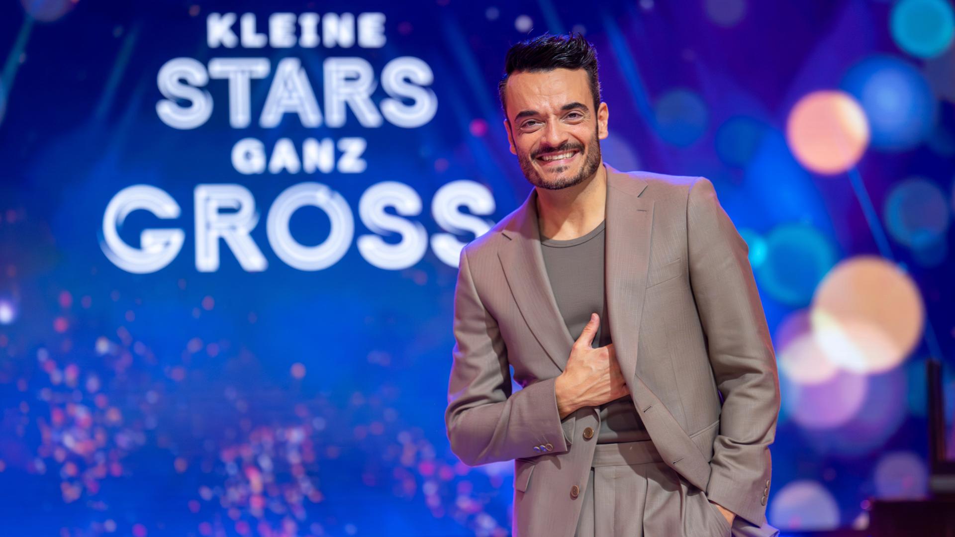 "Die Giovanni Zarrella Show – Kleine Stars ganz groß": Giovanni Zarrella steht im graubraunen Anzug vor dem Schriftzug mit dem Sendungsmotto "Kleine Stars ganz groß". Er schaut lachend in die Kamera.