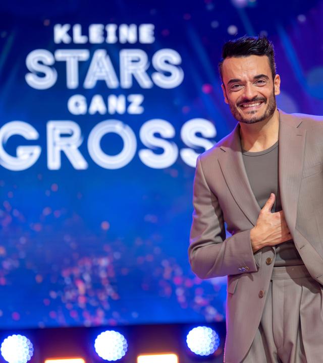 "Die Giovanni Zarrella Show – Kleine Stars ganz groß": Giovanni Zarrella steht im graubraunen Anzug vor dem Schriftzug mit dem Sendungsmotto "Kleine Stars ganz groß". Er schaut lachend in die Kamera.