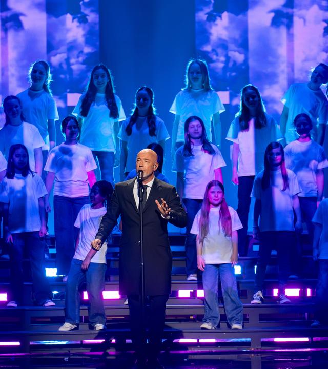 "Die Giovanni Zarrella Show – Kleine Stars ganz groß": Der Graf, Sänger der Band Unheilig, steht auf der Bühne und singt in ein Standmikrofon. Hinter ihm auf einer Showtreppe steht der Kinderchor Lucky Kids.
