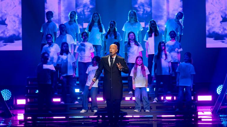 "Die Giovanni Zarrella Show – Kleine Stars ganz groß": Der Graf, Sänger der Band Unheilig, steht auf der Bühne und singt in ein Standmikrofon. Hinter ihm auf einer Showtreppe steht der Kinderchor Lucky Kids.