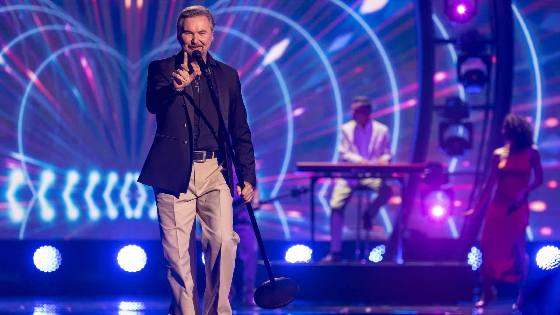 "Die Giovanni Zarrella Show – Kleine Stars ganz groß": Nik P. läuft singend über die Bühne, trägt ein Standmikrofon in der linken Hand und zeigt mit dem rechten Zeigefinger nach oben. Im Hintergrund befinden sich die Musiker der Livebandleuchten.