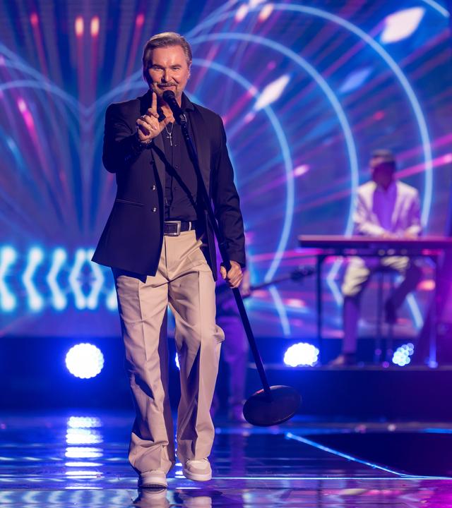 "Die Giovanni Zarrella Show – Kleine Stars ganz groß": Nik P. läuft singend über die Bühne, trägt ein Standmikrofon in der linken Hand und zeigt mit dem rechten Zeigefinger nach oben. Im Hintergrund befinden sich die Musiker der Livebandleuchten.