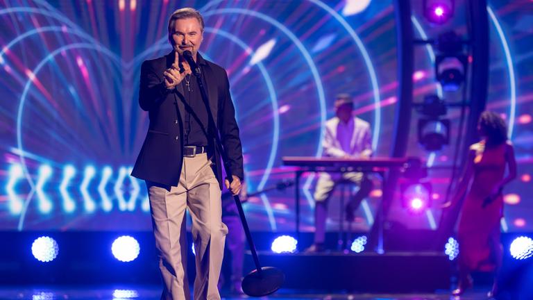 "Die Giovanni Zarrella Show – Kleine Stars ganz groß": Nik P. läuft singend über die Bühne, trägt ein Standmikrofon in der linken Hand und zeigt mit dem rechten Zeigefinger nach oben. Im Hintergrund befinden sich die Musiker der Livebandleuchten.