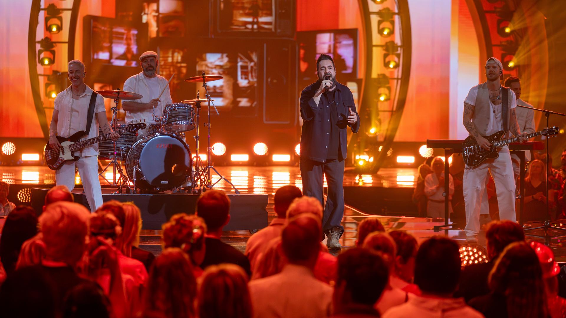 "Die Giovanni Zarrella Show – Kleine Stars ganz groß": Laith Al-Deen steht gemeinsam mit der Band der "Giovanni Zarrella Show" auf der Bühne. Das Licht ist in warmen Orangetönen gestaltet. Im Vordergrund sitzt das Publikum.