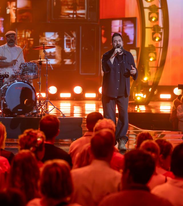 "Die Giovanni Zarrella Show – Kleine Stars ganz groß": Laith Al-Deen steht gemeinsam mit der Band der "Giovanni Zarrella Show" auf der Bühne. Das Licht ist in warmen Orangetönen gestaltet. Im Vordergrund sitzt das Publikum.