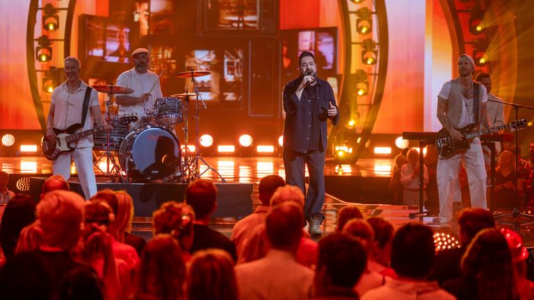 "Die Giovanni Zarrella Show – Kleine Stars ganz groß": Laith Al-Deen steht gemeinsam mit der Band der "Giovanni Zarrella Show" auf der Bühne. Das Licht ist in warmen Orangetönen gestaltet. Im Vordergrund sitzt das Publikum.