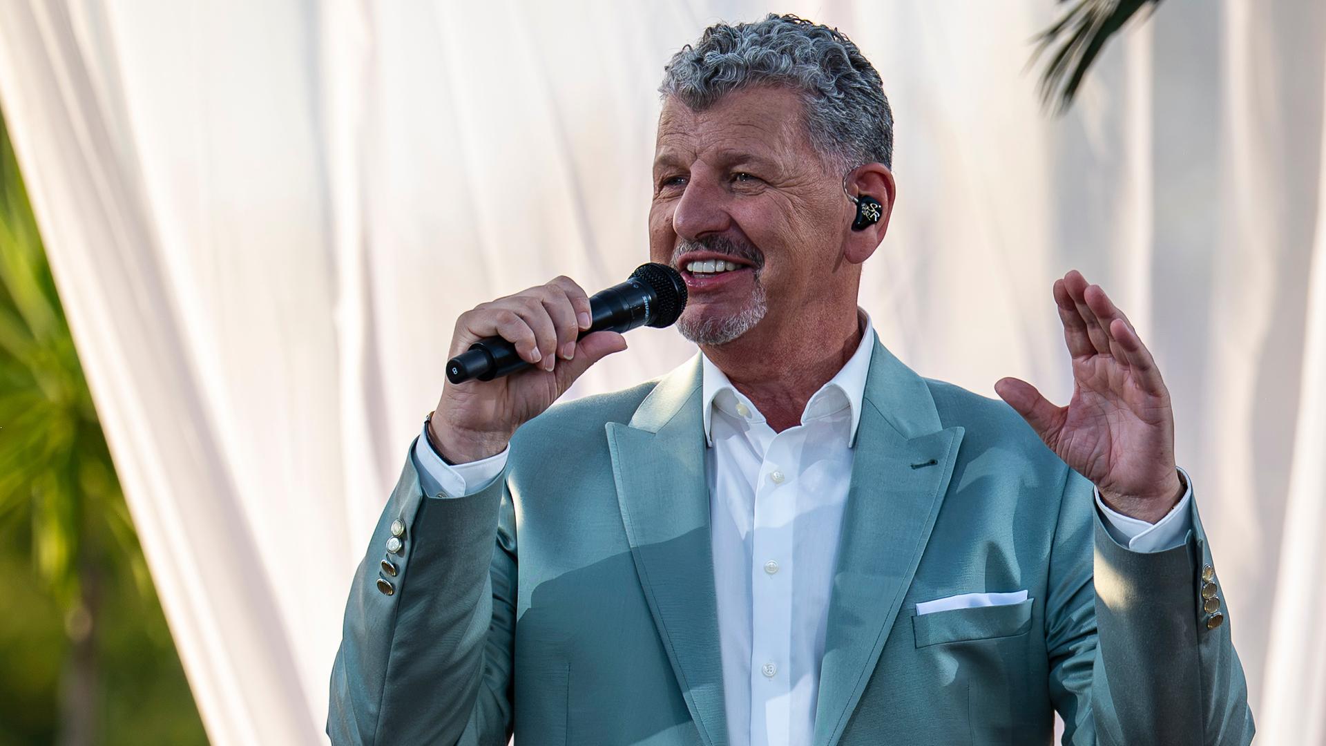 "Die Giovanni Zarrella Show - Die große Sommerparty": Semino Rossi singt lächelnd in ein Handmikrofon. Seine linke Hand zeigt nach oben. Im Hintergrund stehen Palmen hinter einem weißen Vorhang. Er trägt einen eleganten petrolfarbenen Anzug und ein weißes Hemd.