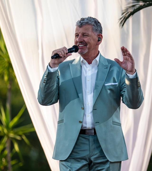 "Die Giovanni Zarrella Show - Die große Sommerparty": Semino Rossi singt lächelnd in ein Handmikrofon. Seine linke Hand zeigt nach oben. Im Hintergrund stehen Palmen hinter einem weißen Vorhang. Er trägt einen eleganten petrolfarbenen Anzug und ein weißes Hemd.