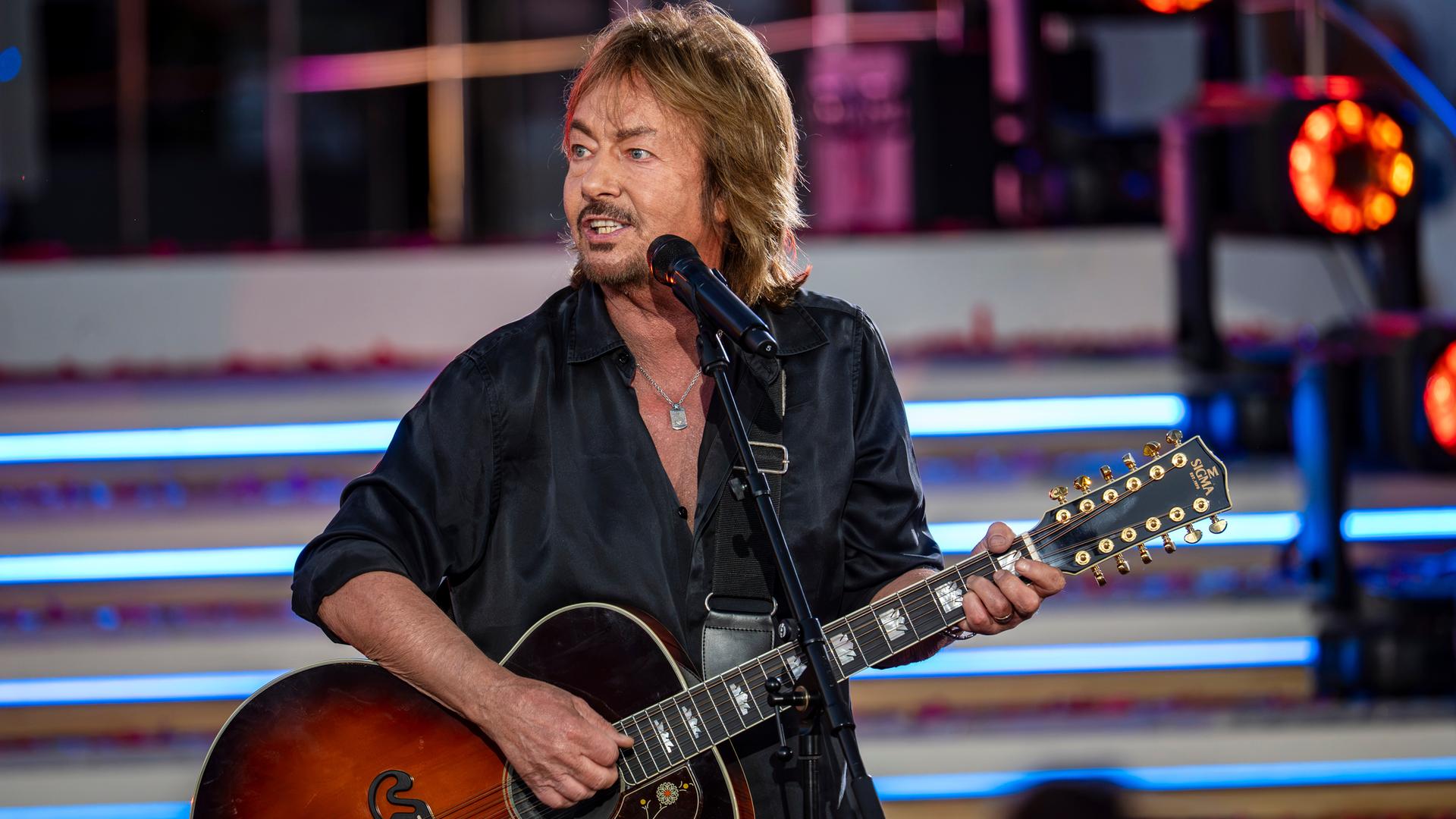 "Die Giovanni Zarrella Show - Die große Sommerparty": Chris Norman steht mit einer Akustikgitarre an einem Standmikrofon auf der Bühne. Er trägt ein schwarzes Hemd und blickt zur Seite. Im Hintergrund beleuchten Scheinwerfer die Showtreppe.