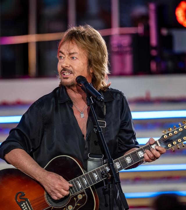 "Die Giovanni Zarrella Show - Die große Sommerparty": Chris Norman steht mit einer Akustikgitarre an einem Standmikrofon auf der Bühne. Er trägt ein schwarzes Hemd und blickt zur Seite. Im Hintergrund beleuchten Scheinwerfer die Showtreppe.