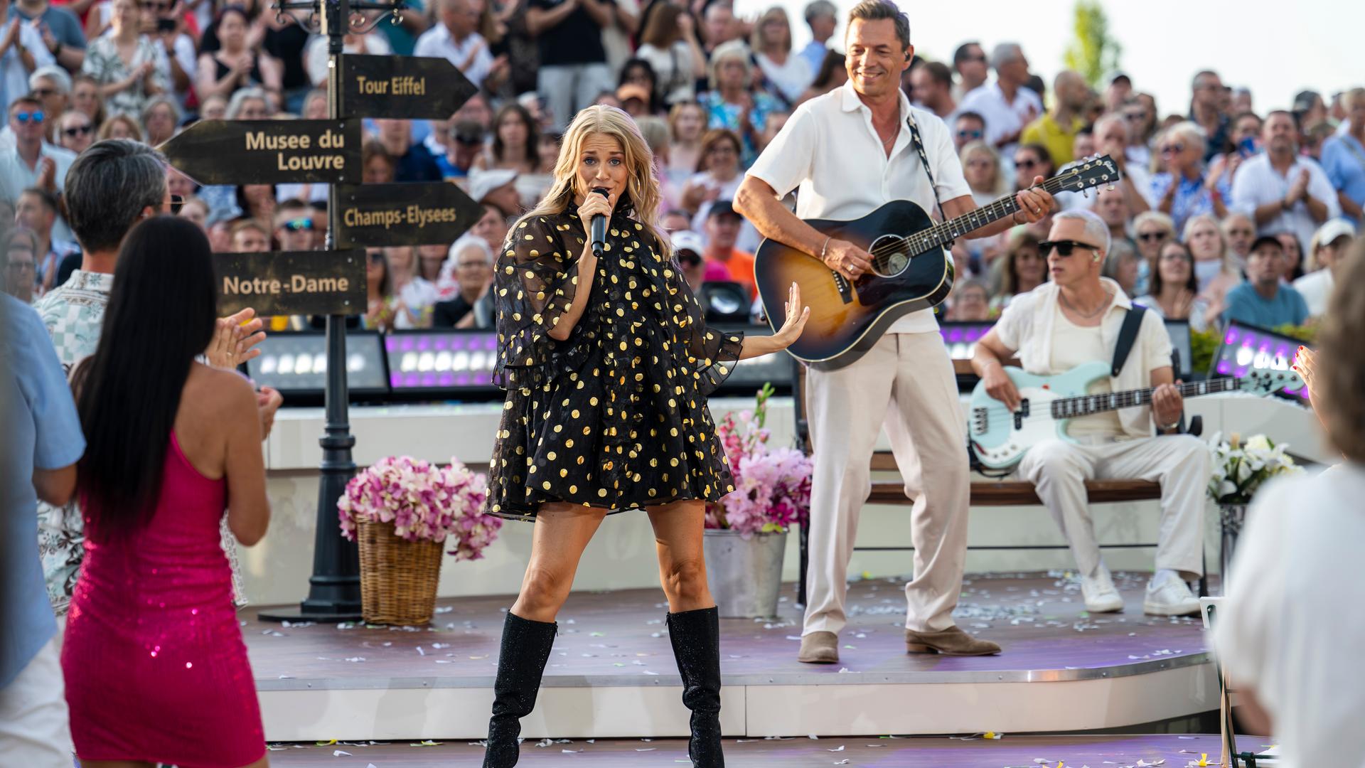 "Die Giovanni Zarrella Show - Die große Sommerparty": Julia Lindholm und der Gitarrist und Bassist der Live-Band stehen auf der Bühne. Sie singt und ist umgeben von Publikum. Die Bühne ist dekoriert mit einer Laterne und Hinweisschildern von Pariser Sehenswürdigkeiten.
