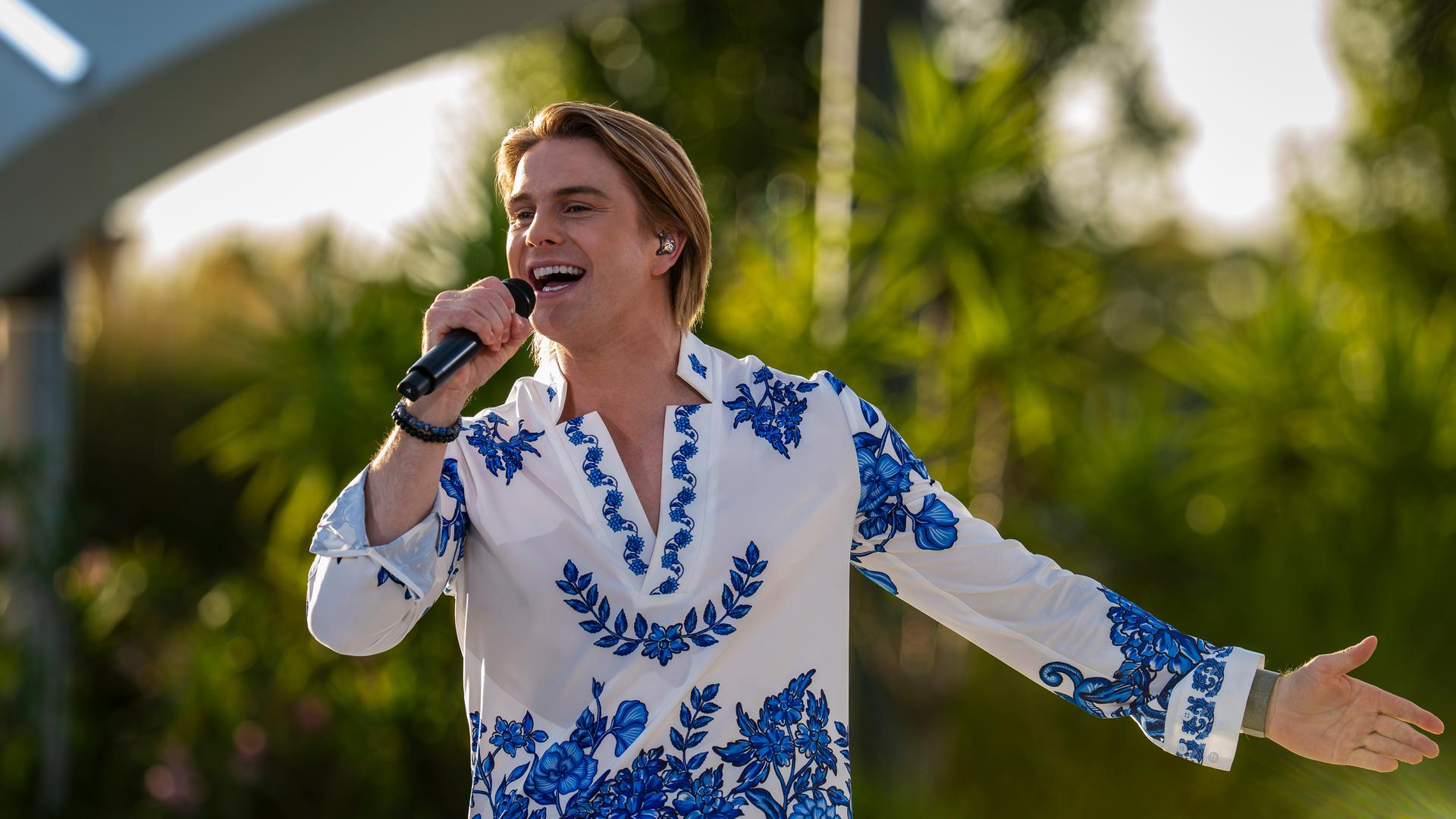 "Die Giovanni Zarrella Show - Die große Sommerparty": Schlagerstar Vincent Gross singt gut gelaunt in ein Handmikrofon. Die linke Hand zeigt zur Seite. Er trägt ein weißes Hemd mit blauen floralen Motiven. Im Hintergrund in der Unschärfe befinden sich Bäume und Palmen.