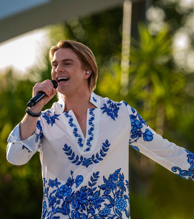 "Die Giovanni Zarrella Show - Die große Sommerparty": Schlagerstar Vincent Gross singt gut gelaunt in ein Handmikrofon. Die linke Hand zeigt zur Seite. Er trägt ein weißes Hemd mit blauen floralen Motiven. Im Hintergrund in der Unschärfe befinden sich Bäume und Palmen.