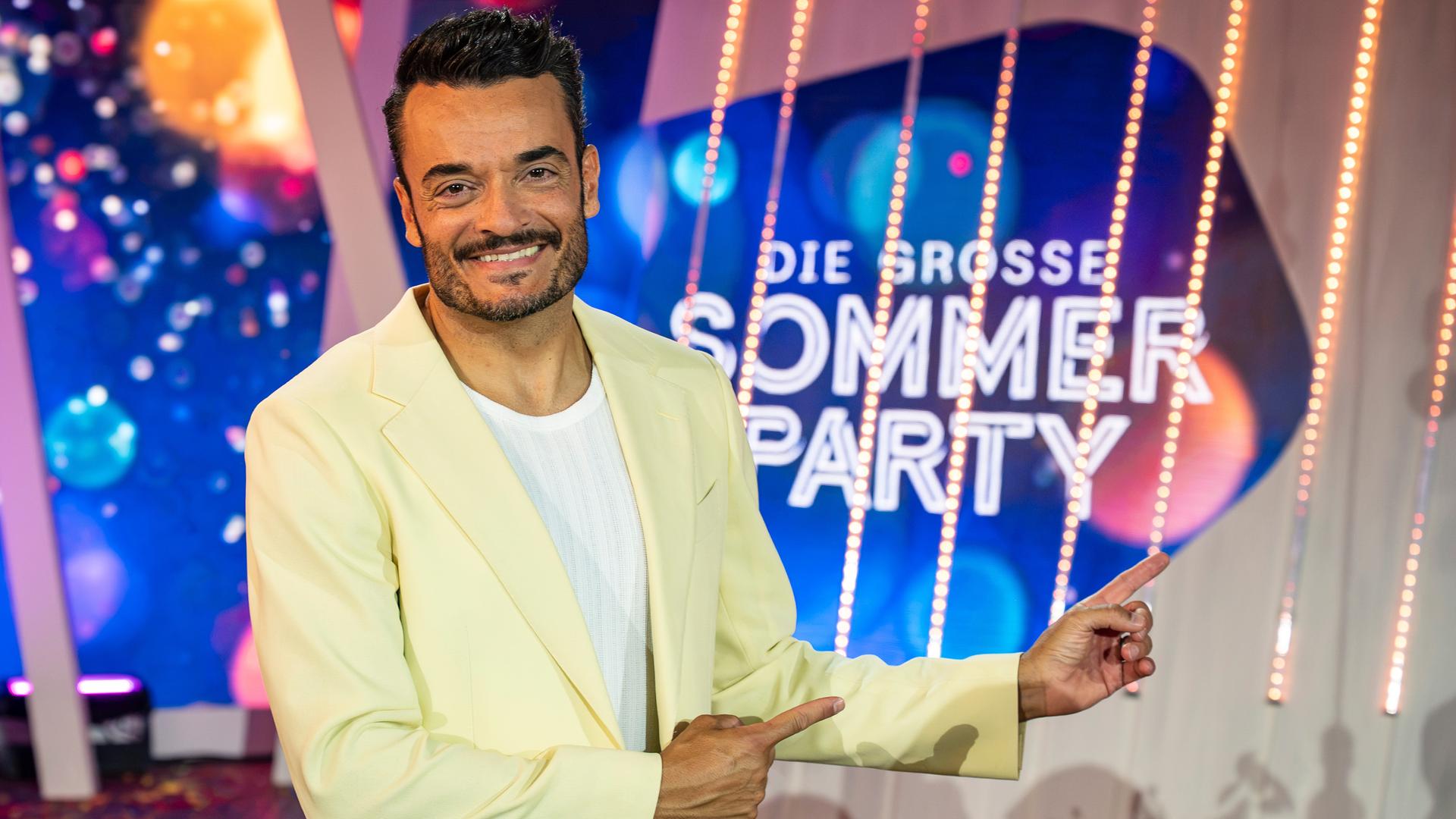 "Die Giovanni Zarrella Show - Die große Sommerparty": Moderator Giovanni Zarrella lächelt in die Kamera und zeigt mit beiden Zeigefingern auf das Sendungslogo "Die große Sommerparty". Er trägt einen cremefarbenen Anzug und ein weißes T-Shirt.