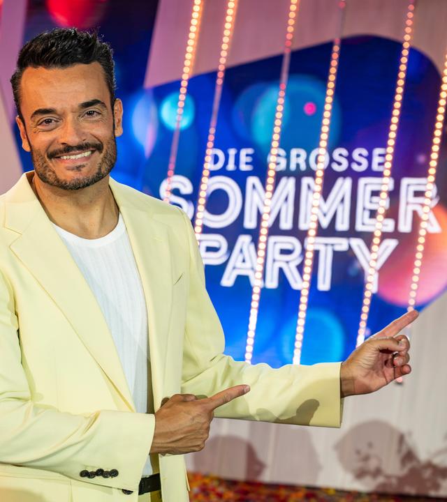 "Die Giovanni Zarrella Show - Die große Sommerparty": Moderator Giovanni Zarrella lächelt in die Kamera und zeigt mit beiden Zeigefingern auf das Sendungslogo "Die große Sommerparty". Er trägt einen cremefarbenen Anzug und ein weißes T-Shirt.