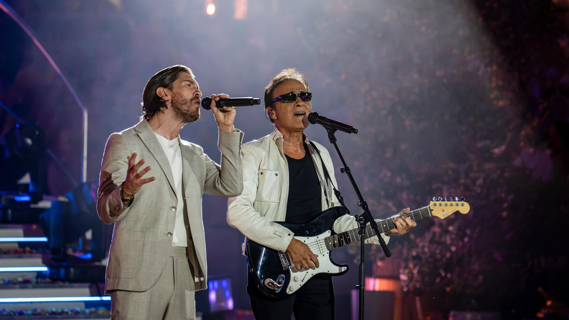 "Die Giovanni Zarrella Show - Die große Sommerparty": Joey Heindle und F. R. David stehen auf der Bühne und singen im Duett. Joey Heindle trägt einen hellen Sommeranzug und singt in ein Handmikrofon. Neben ihm steht F.R. David mit weißer Lederjacke und Sonnenbrille. Er spielt E-Gitarre und singt in ein Standmikrofon. Der Hintergrund ist dunkel, das Publikum ist schemenhaft zu erkennen.