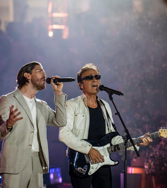 "Die Giovanni Zarrella Show - Die große Sommerparty": Joey Heindle und F. R. David stehen auf der Bühne und singen im Duett. Joey Heindle trägt einen hellen Sommeranzug und singt in ein Handmikrofon. Neben ihm steht F.R. David mit weißer Lederjacke und Sonnenbrille. Er spielt E-Gitarre und singt in ein Standmikrofon. Der Hintergrund ist dunkel, das Publikum ist schemenhaft zu erkennen.