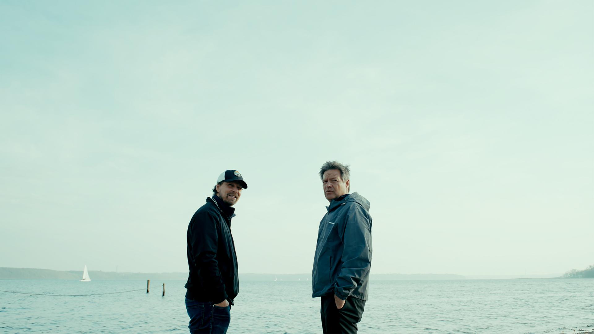 Zwei Männer in Windbreakern stehen an einem Strand.