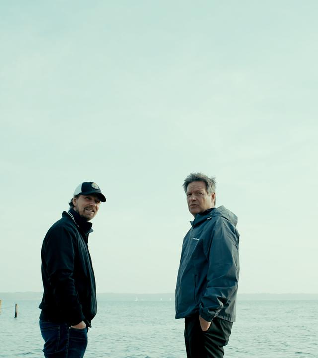 Zwei Männer in Windbreakern stehen an einem Strand.